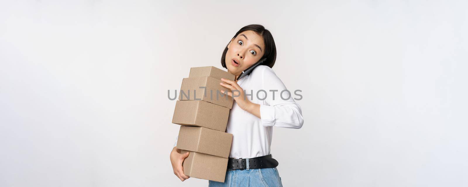Young asian businesswoman answer phone call, talking on mobile while carrying pile of boxes with orders, standing over white background by Benzoix
