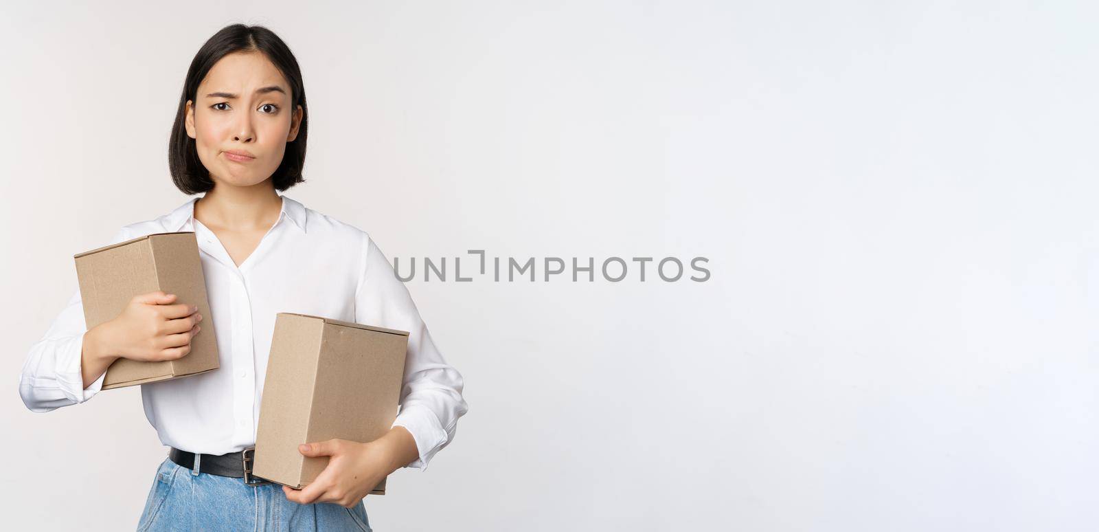 Complicated young asian woman holding two boxes, looking doubtful at camera, standing over white background puzzled by Benzoix