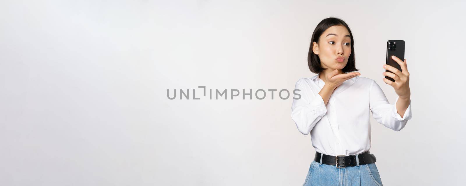 Image of asian cute girl video chat, sending air kiss at camera, taking selfie with app filters on smartphone, standing over white background.