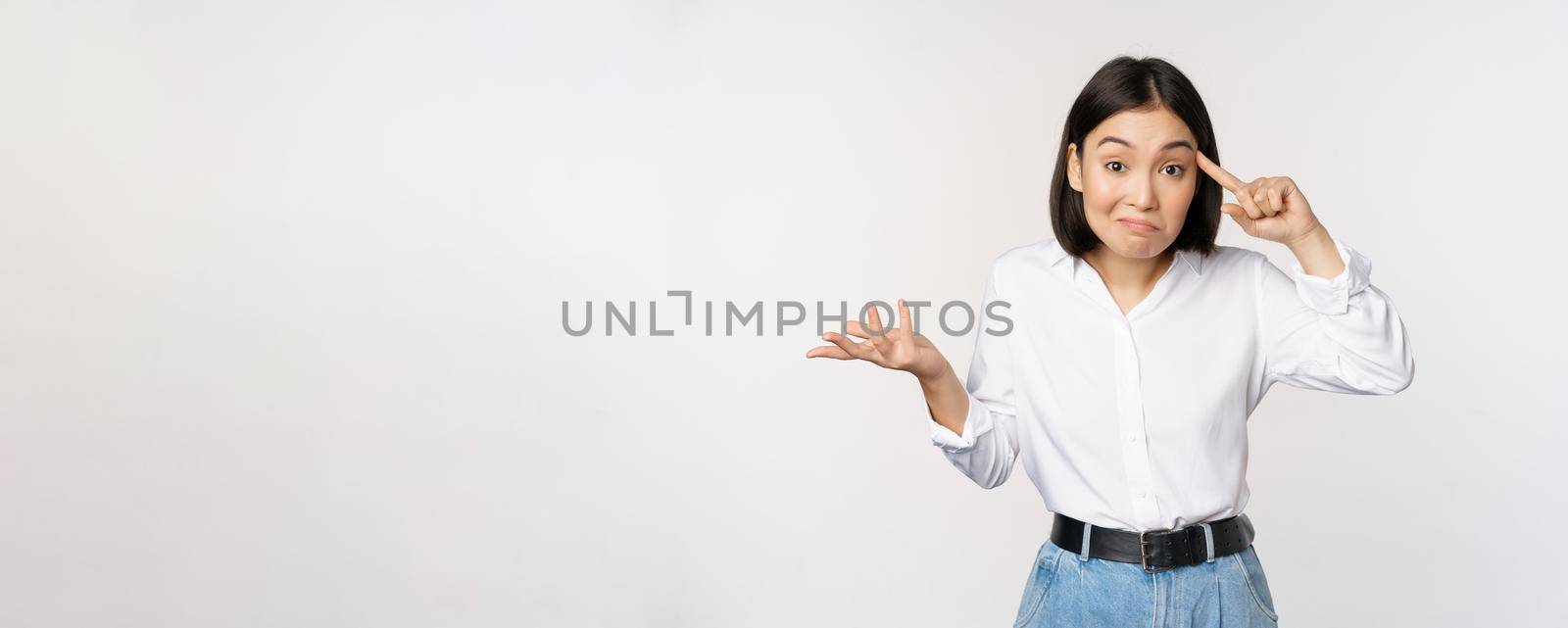 Are you insane. Portrait of frustrated young asian woman, roll finger near head and srugging, someone crazy or stupid, standing over white background by Benzoix