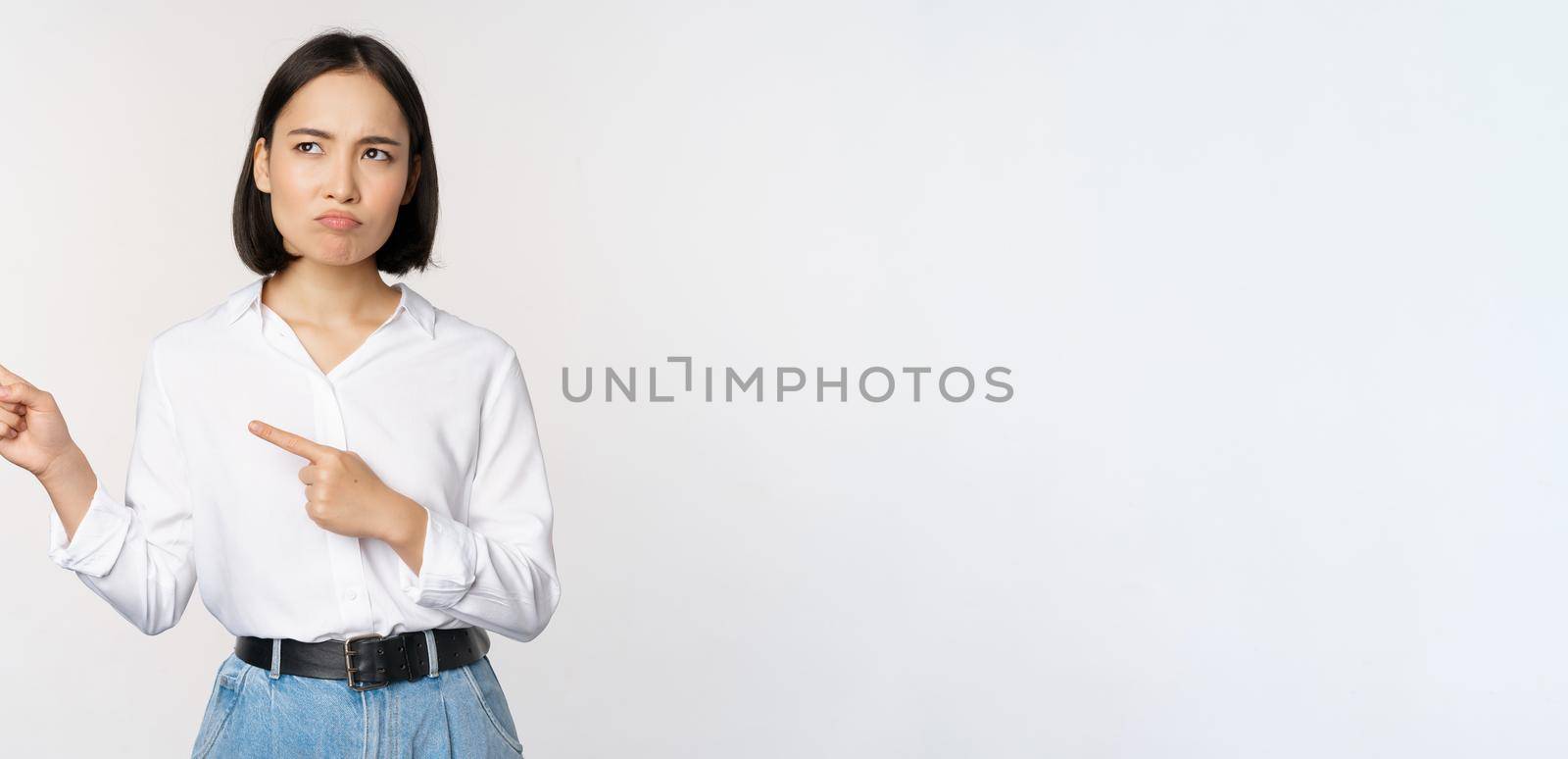 Disappointed, sad young asian woman pointing and looking left with upset, sulking face expression, standing over white background.