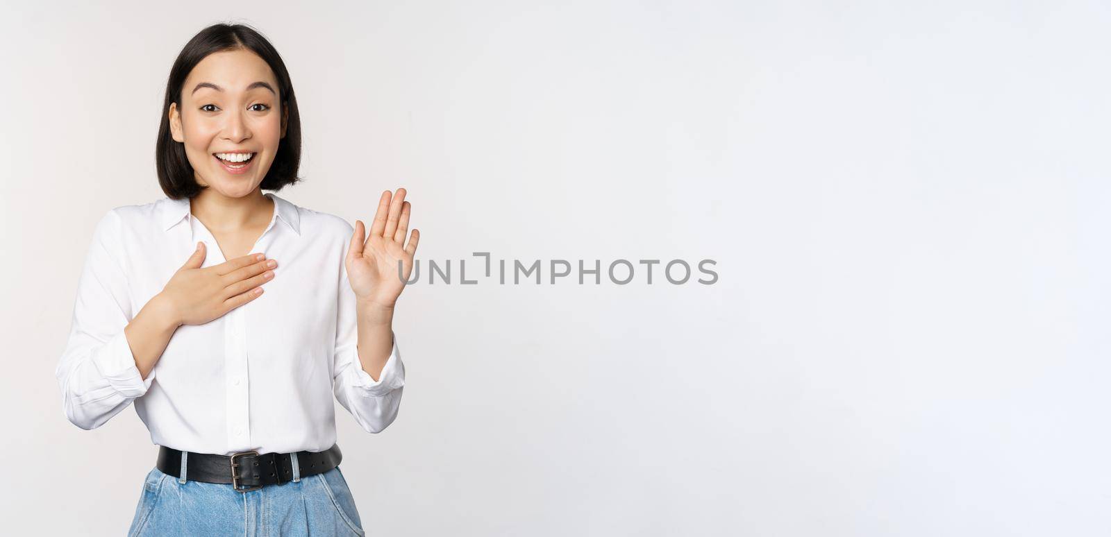 Image of cute young female office worker, asian girl student raising hand up and put palm on chest, name herself, introduce, making promise, standing over white background.