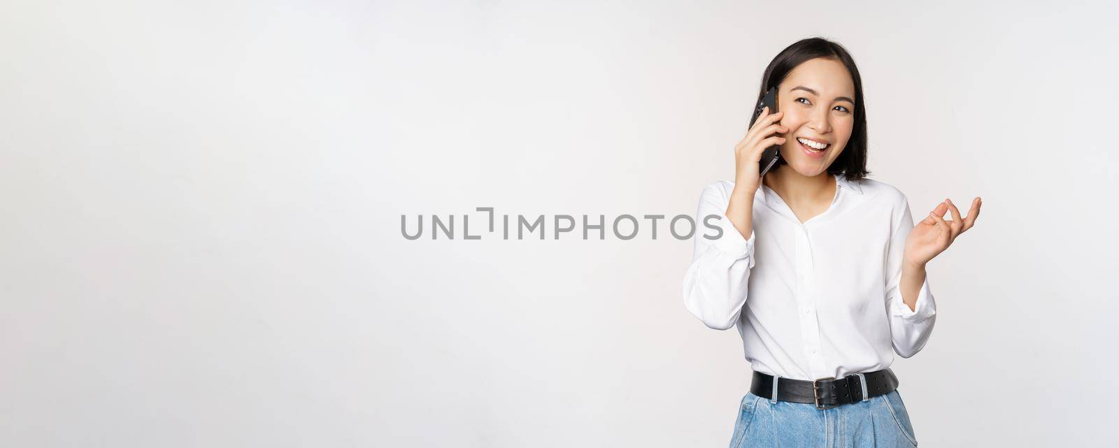 Friendly smiling asian woman talking on phone, girl on call, holding smartphone and laughing, speaking, standing over white background by Benzoix