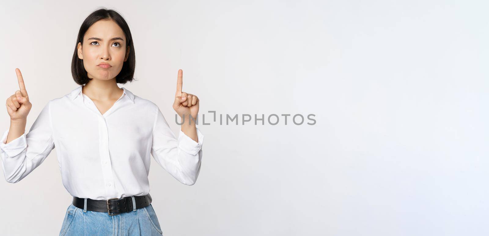 Skeptical businesswoman, asian office manager, pointing fingers up and grimacing doubtful, hesitating, standing over white background.