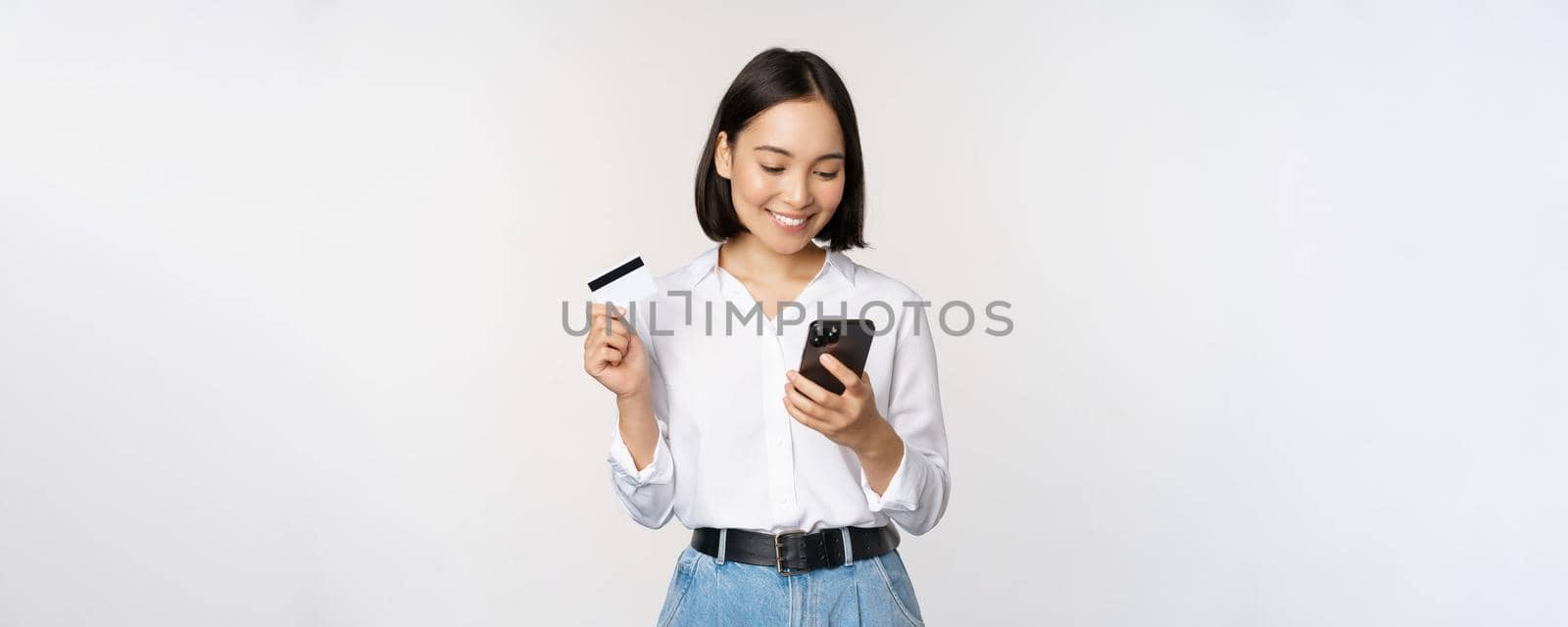 Online shopping concept. Korean woman holding credit card and looking at smartphone app, buying, order delivery in mobile phone application, standing over white background by Benzoix