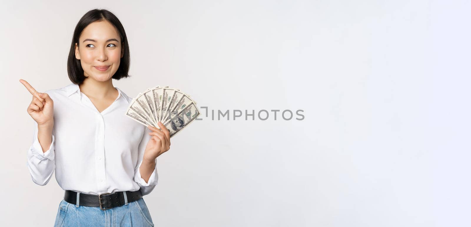 Smiling young modern asian woman, pointing at banner advertisement, holding cash money dollars, standing over white background.