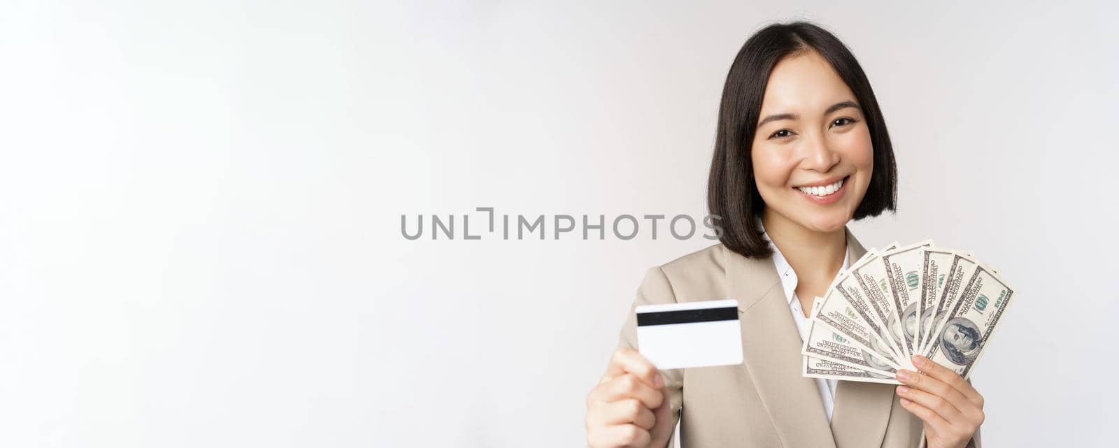 Close up of asian businesswoman, office lady showing credit card and money dollars, standing in suit over white background.