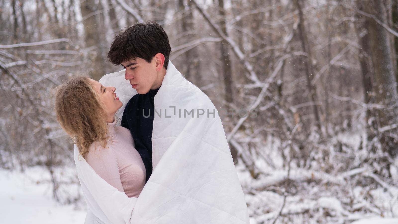 A young couple walks in the park in winter. The guy and the girl are kissing wrapped in a white blanket outdoors