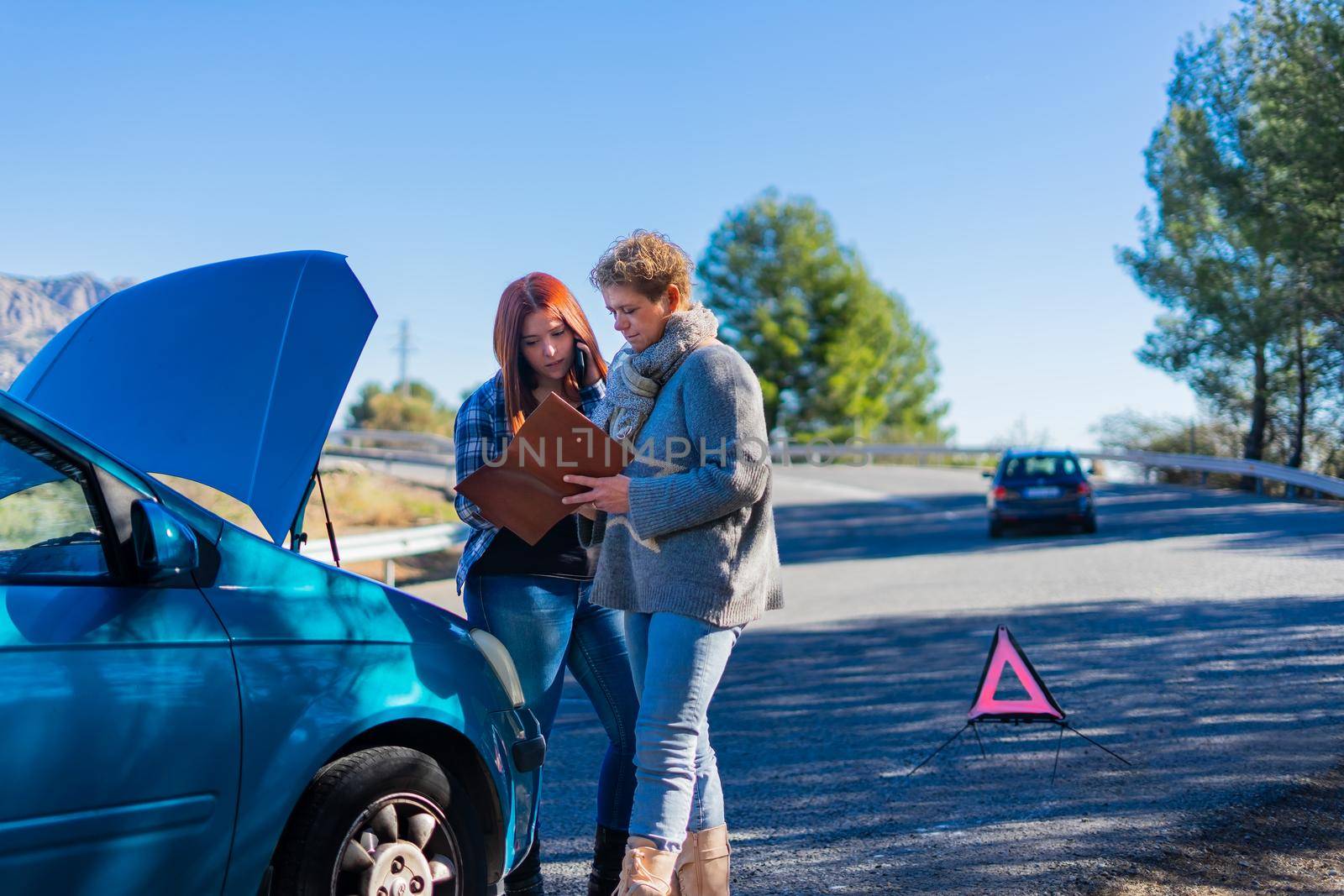 Mother and daughter standing next to the broken down vehicle checking the documents and calling the tow truck. Two friends on holiday have a car accident and have to call the mechanic in the middle of the road. Clear road on a very sunny day next to a forest with a mountain in the background. Red-haired girl calling tow truck and blonde woman looking at insurance papers.