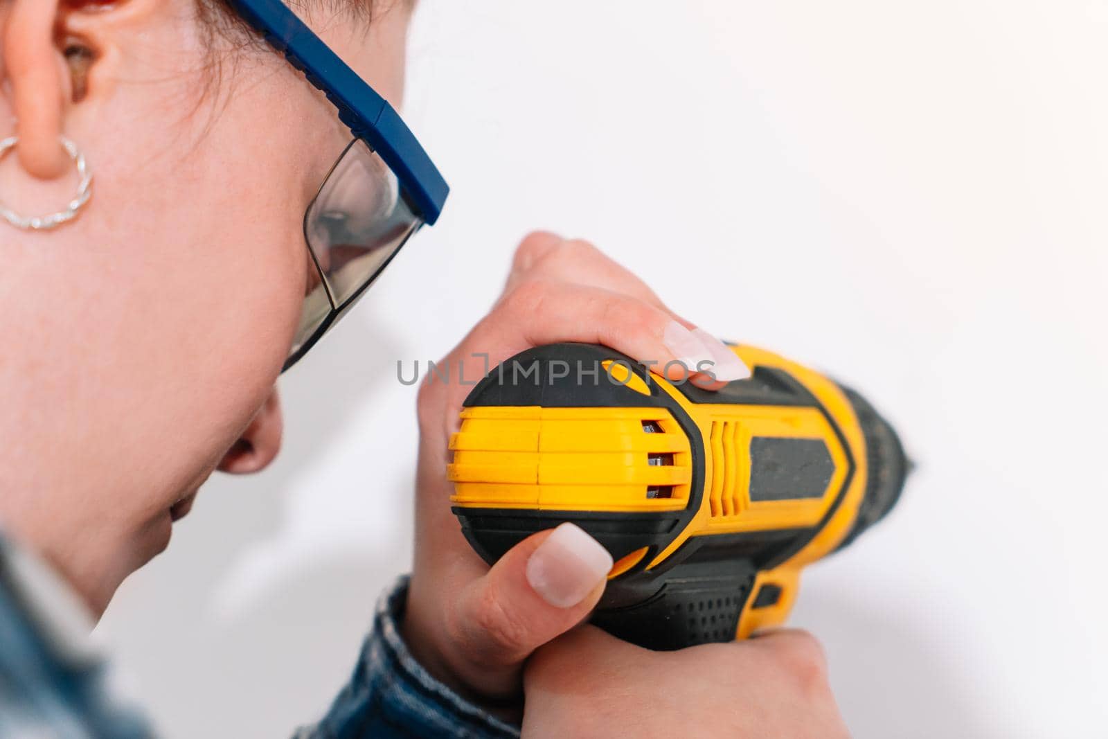 Young girl with red hair making a hole in a white wall with a drill to place a picture or a shelf. The woman is wearing blue safety goggles, a blue and white checked shirt and a yellow battery-powered drill. Her hair is tied back.