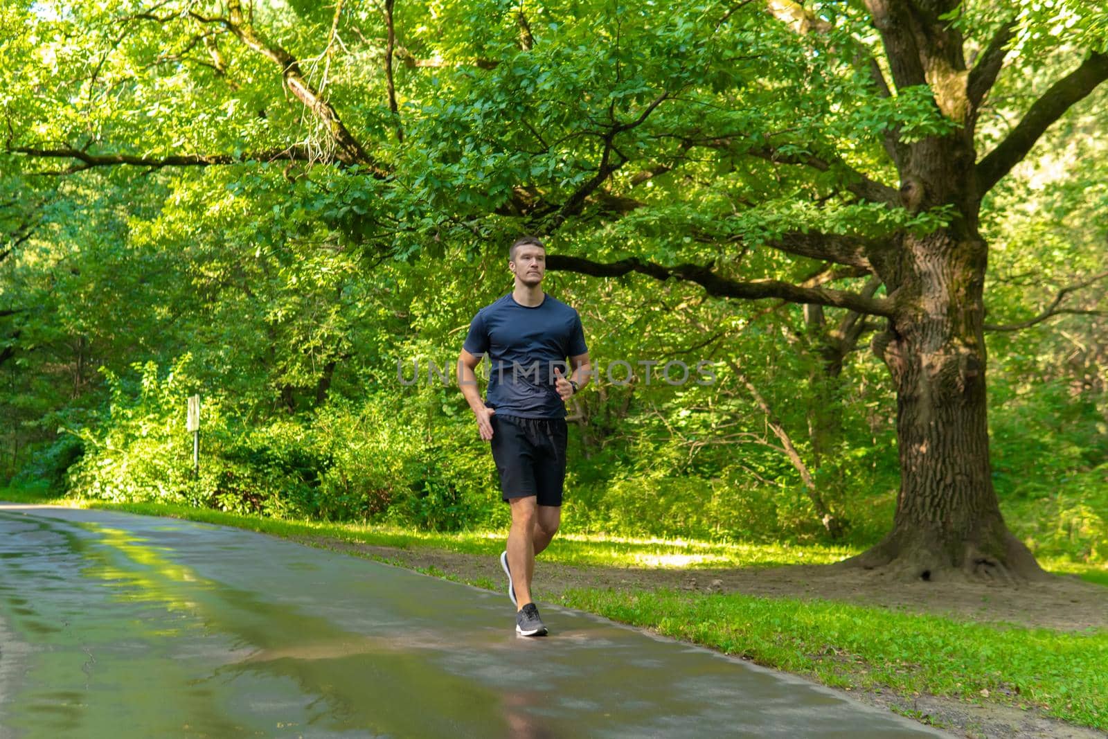 A man athlete runs in the park outdoors, around the forest, oak trees green grass young enduring athletic athlete healthy exercise, fitness trail training legs male, woods outside. Summer leisure running, runners stretches