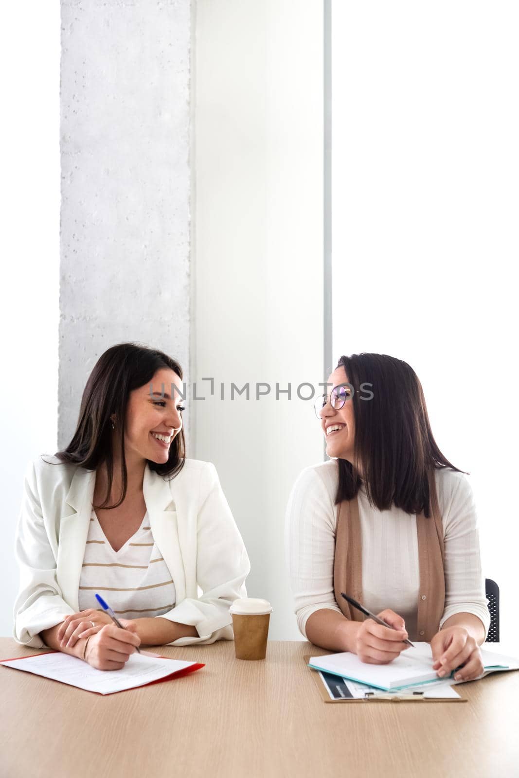 Vertical portrait of two female coworkers looking at each other in the office. Copy space. by Hoverstock