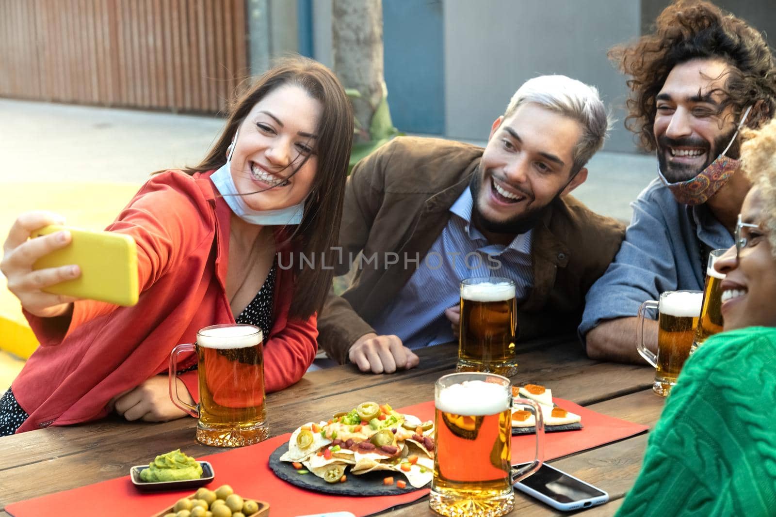 Group of multiracial young friends take selfie with mobile phone in a bar drinking beer. Face mask on chin. Friendship concept.