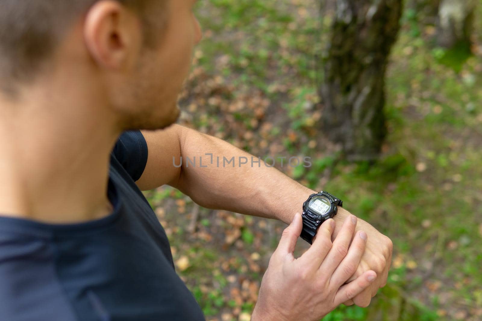 A man looks at the clock time in the park outdoors, around the forest, oaks green grass young enduring athletic athlete. healthy runner fitness trail training person outside. energy stretching stretches