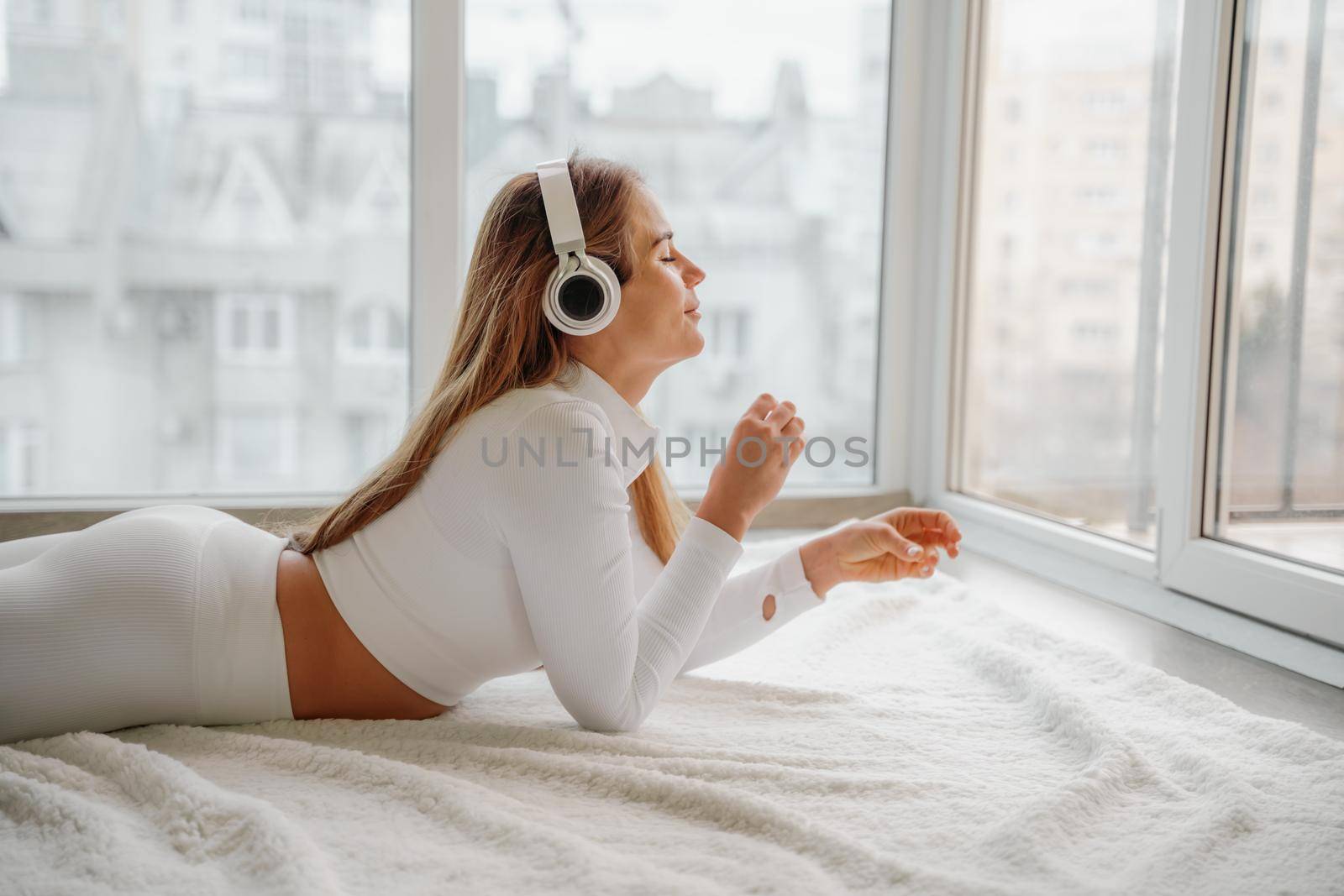 Side view portrait of relaxed woman listening to music with headphones lying on carpet at home. She is dressed in a white tracksuit