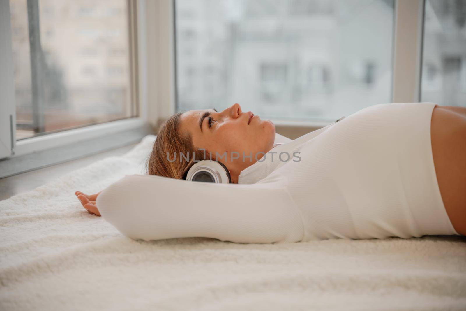 Side view portrait of relaxed woman listening to music with headphones lying on carpet at home. She is dressed in a white tracksuit. by Matiunina