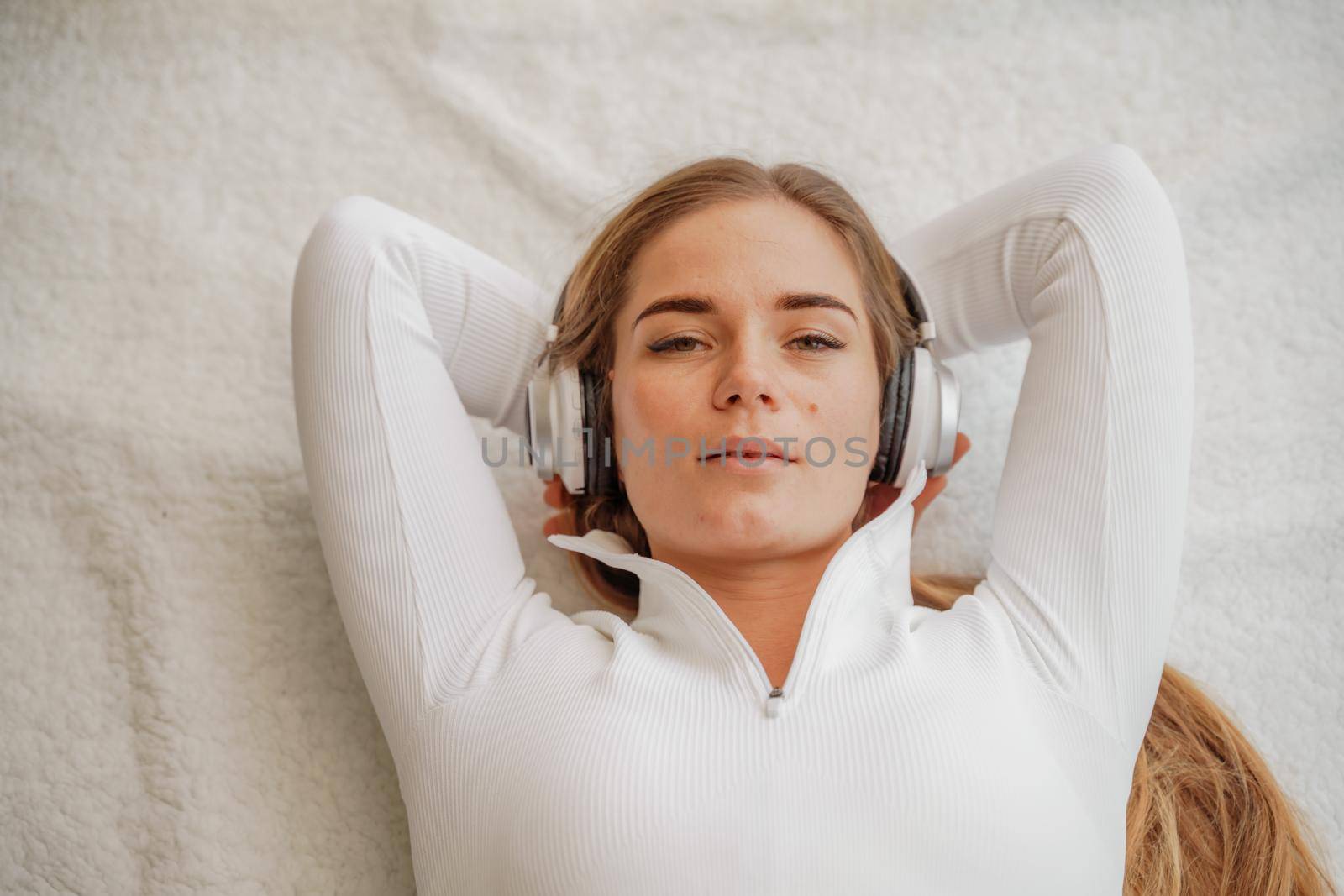 Top view portrait of relaxed woman listening to music with headphones lying on carpet at home. She is dressed in a White tracksuit