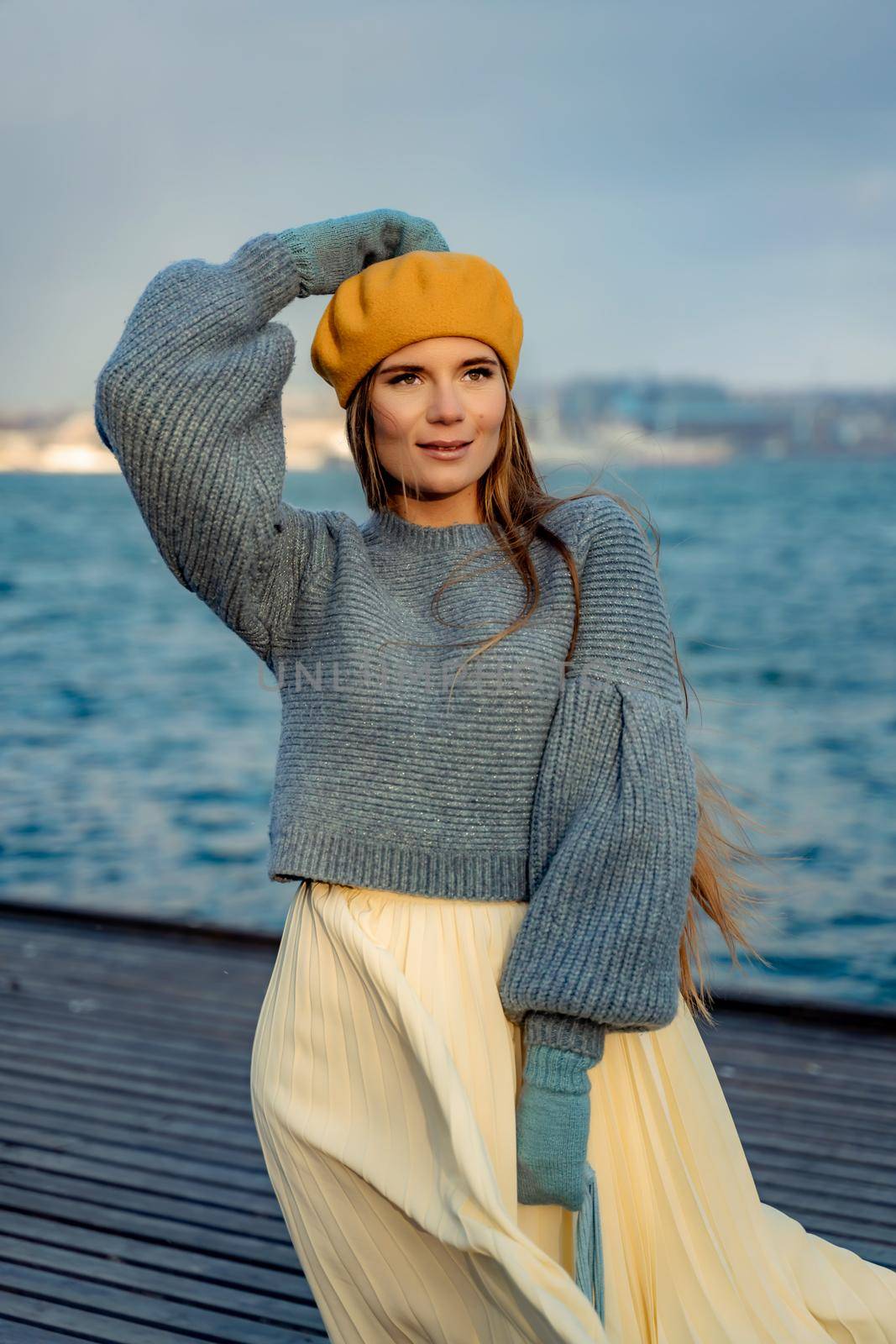 Outdoors fashion portrait of a beautiful middle aged woman walking on the beach. Marine background. Dressed in a stylish warm blue sweater, yellow skirt and beret