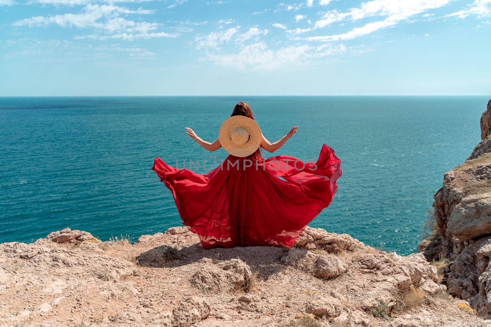 A woman, rear view in a red flying dress fluttering in the wind, a girl in a fluttering dress on the background of the sea. A straw hat hangs at the back of the neck. by Matiunina