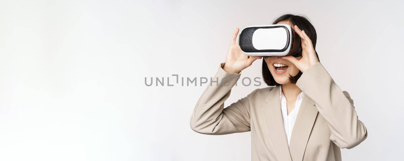 Amazed business woman in suit using virtual reality glasses, looking amazed in vr headset, standing over white background.