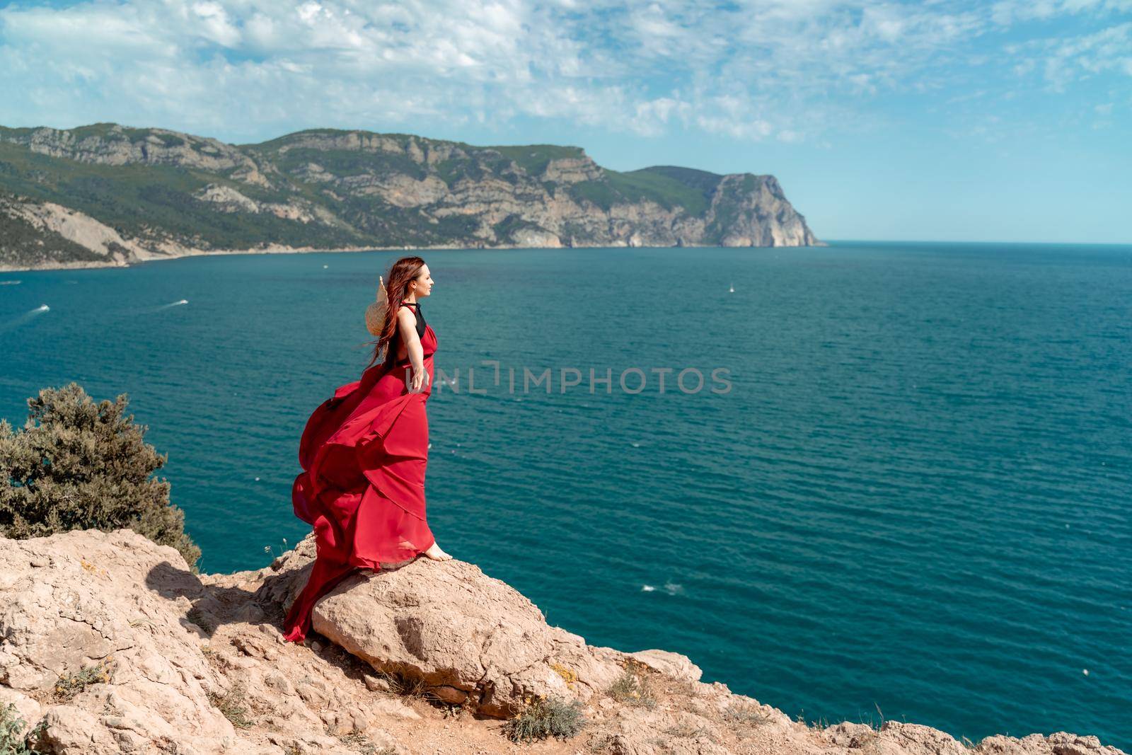 A woman, a side view in a red flying dress fluttering in the wind, a girl in a fluttering dress on the background of the sea. A straw hat hangs at the back of the neck. The concept of a vacation at sea, a photo shoot by the sea, relaxation