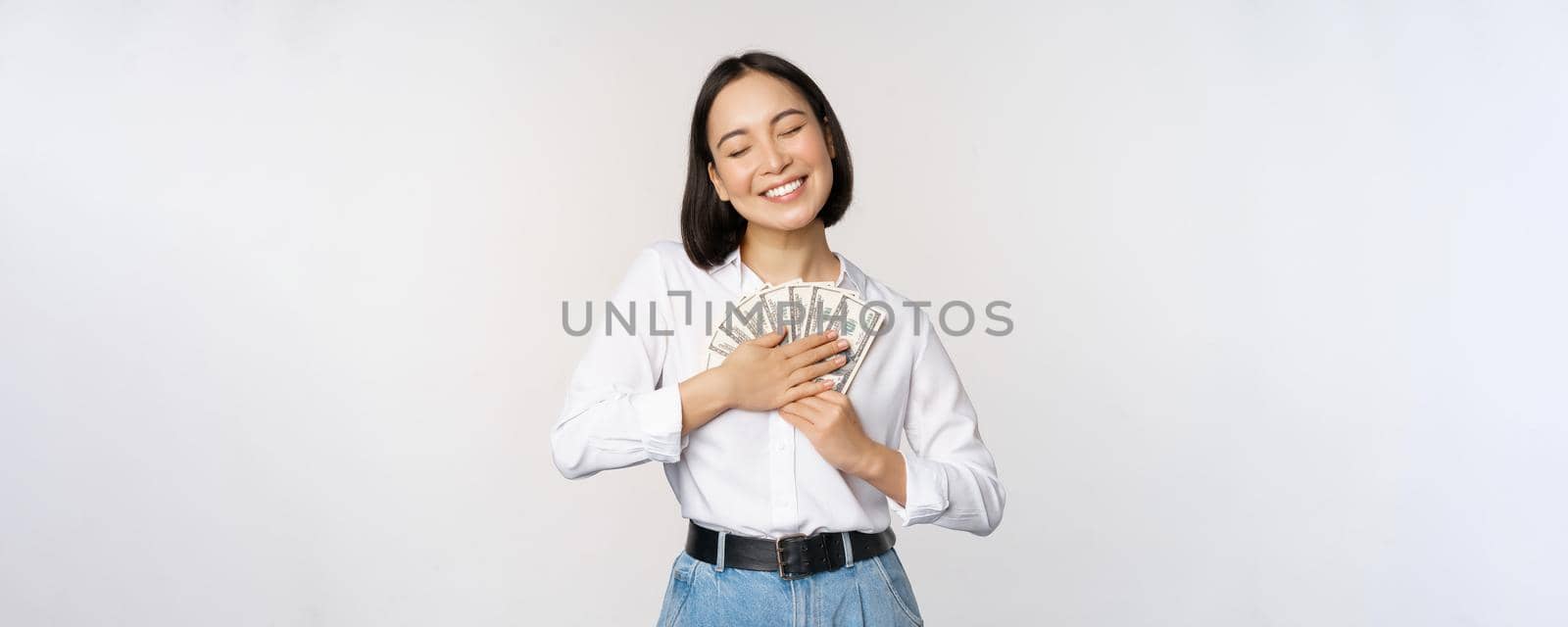 Happy asian woman hugging money dollars and smiling satisfied, standing over white background. Copy space