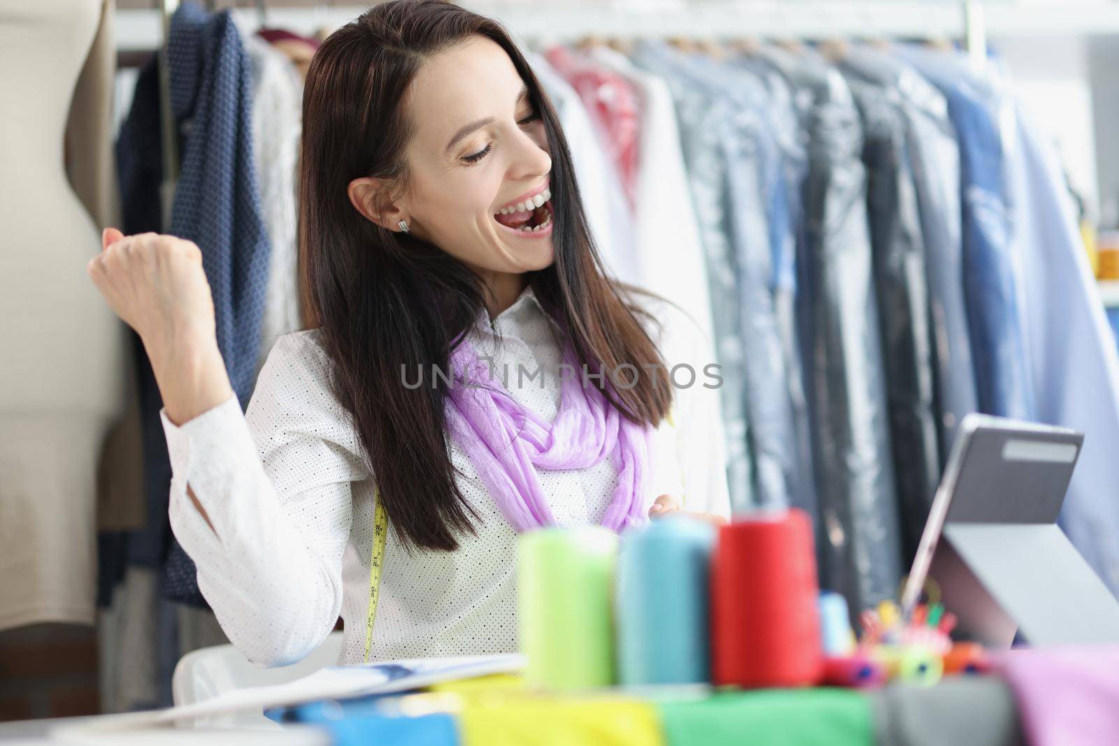 Portrait of happy celebrating dressmaker woman, got beneficial deal, creative showroom. Laughing seamstress on video call. Fashion, career, success concept