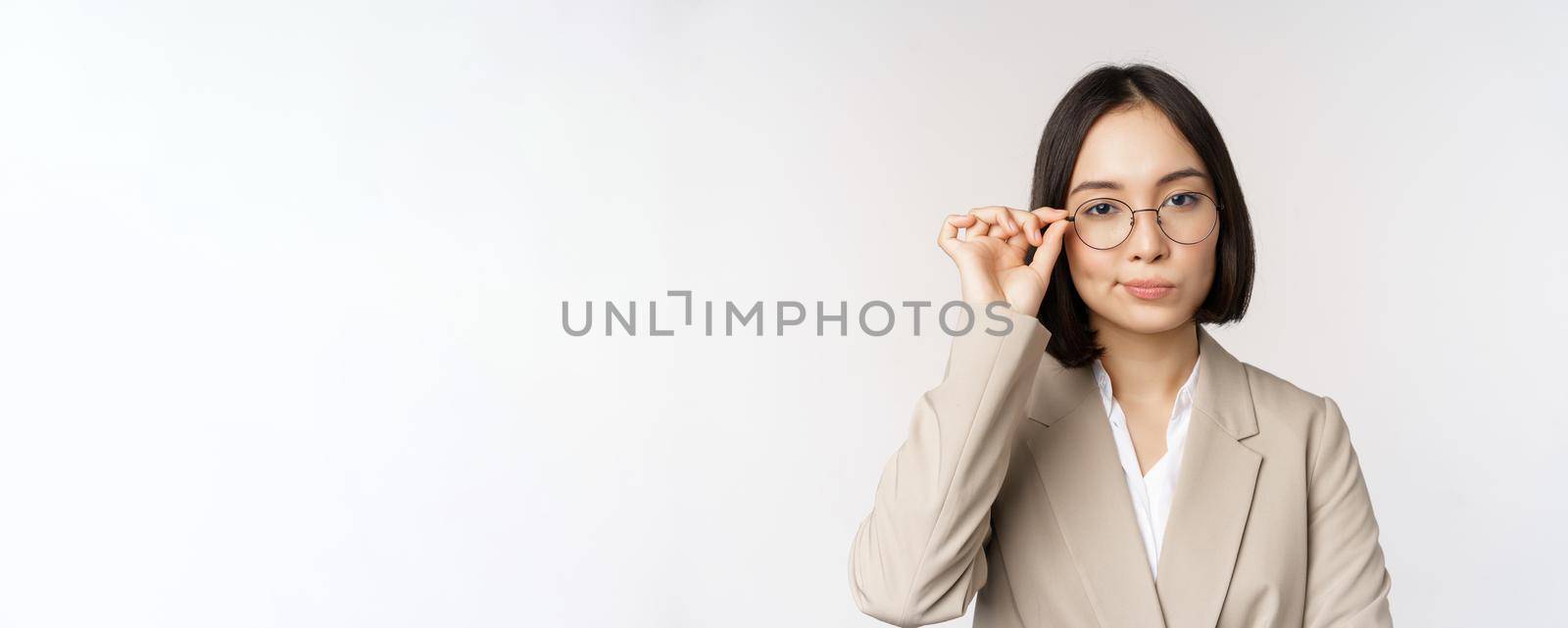 Professional asian businesswoman in glasses, looking confident at camera, standing in power pose against white background.
