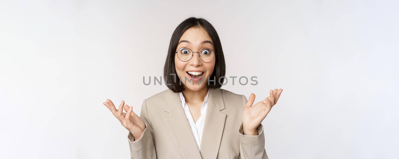 Enthusiastic asian saleswoman in glasses, smiling and laughing, looking amazed at camera, standing in beige suit over white background by Benzoix