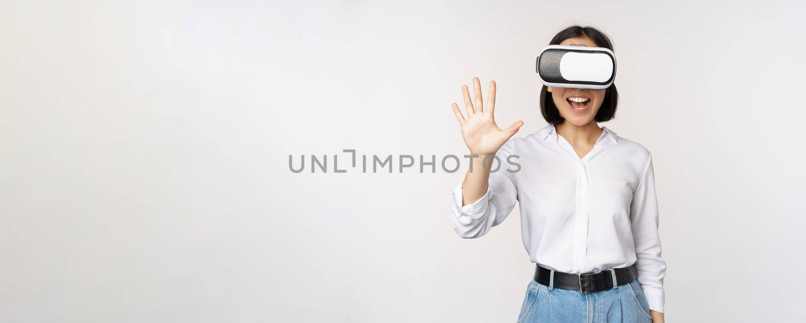 Smiling asian woman waving at you, wearing virtual reality glasses, vr headset, chatting in meta universe, standing over white background.
