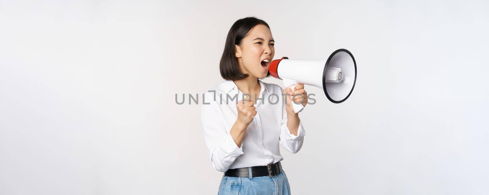 Enthusiastic asian woman, girl activist shouting at protest, using megaphone, looking confident, talking in loudspeaker, protesting, standing over white background by Benzoix