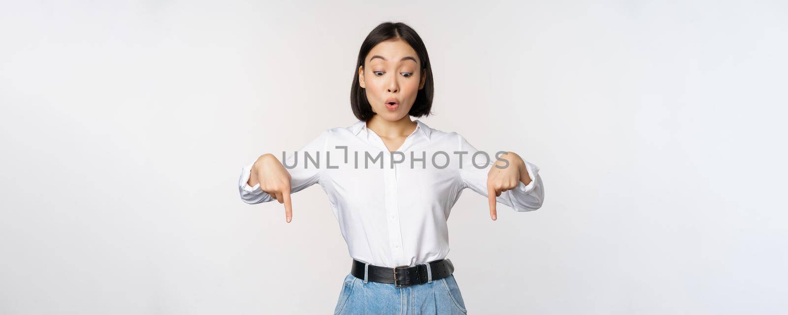 Surprised young asian female student, pointing fingers down and looking with amazed, impressed face expression, standing over white background by Benzoix