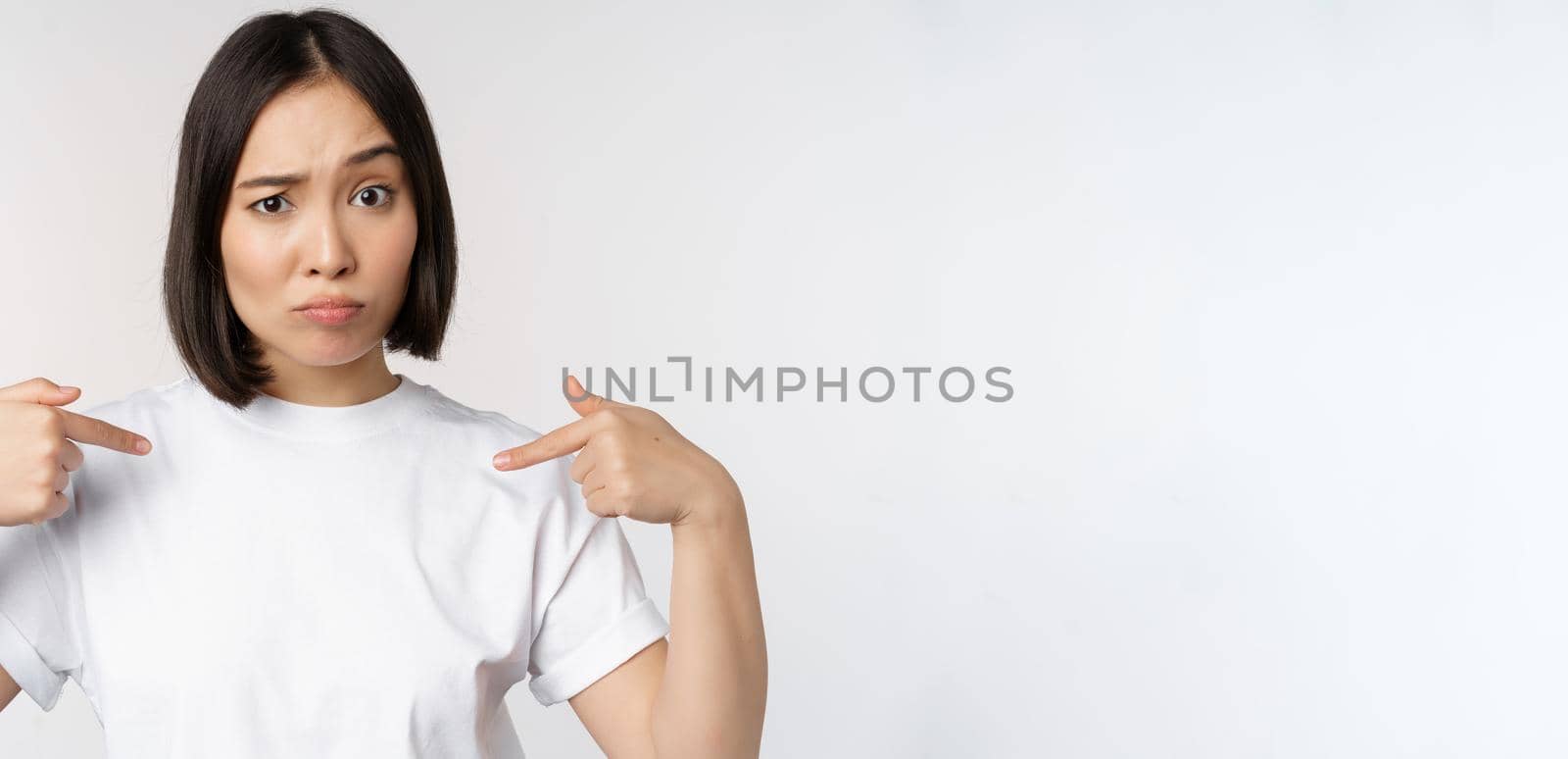 Young asian woman pointing at herself with disbelief, being chosen, surprised by her candidature, standing over white background by Benzoix