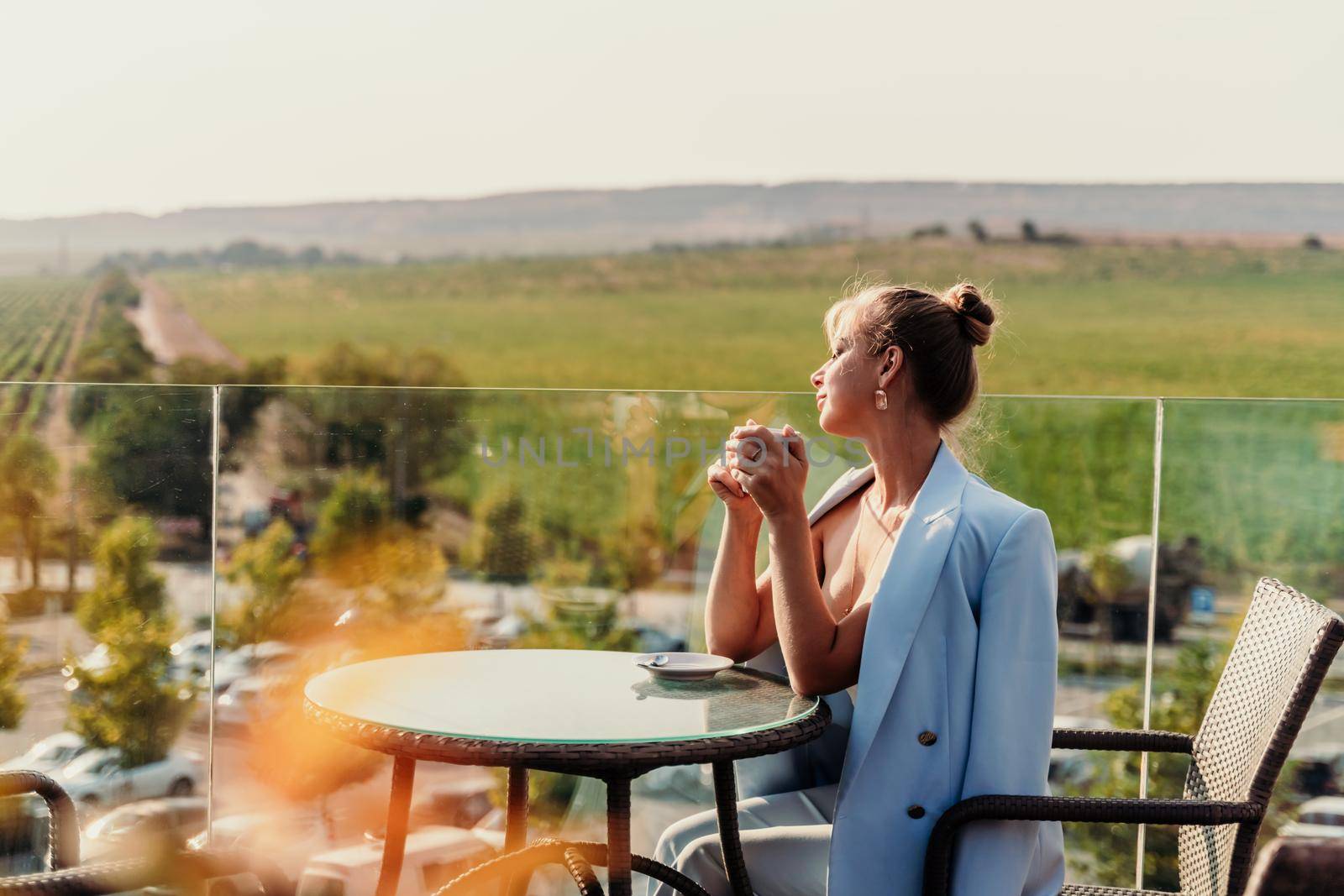 A middle-aged woman sits in a street cafe overlooking the mountains at sunset. She is dressed in a blue jacket and drinks coffee admiring the nature. Travel and vacation concept. by Matiunina