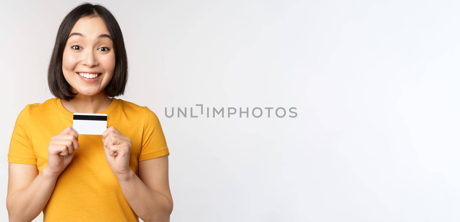 Portrait of beautiful korean girl holding credit card, recommending bank service, standing in yellow tshirt over white background.