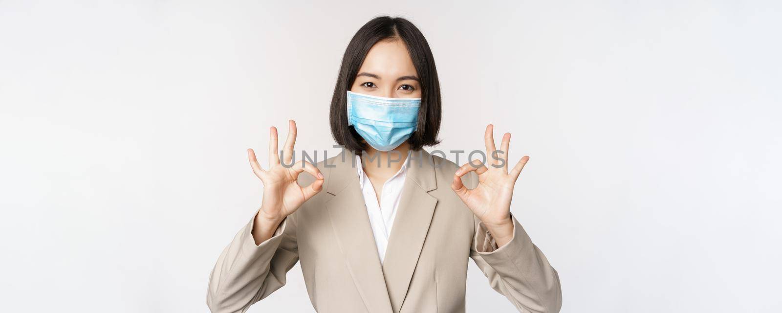 Coronavirus and workplace concept. Image of asian saleswoman, company worker in medical mask showing okay sign, smiling pleased, white background by Benzoix