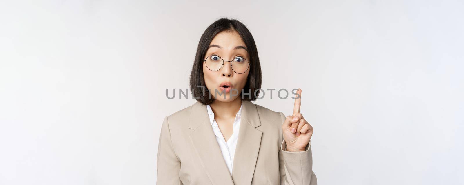 Excited asian woman in glasses, raising finger, eureka sign, has an idea, standing over white background by Benzoix