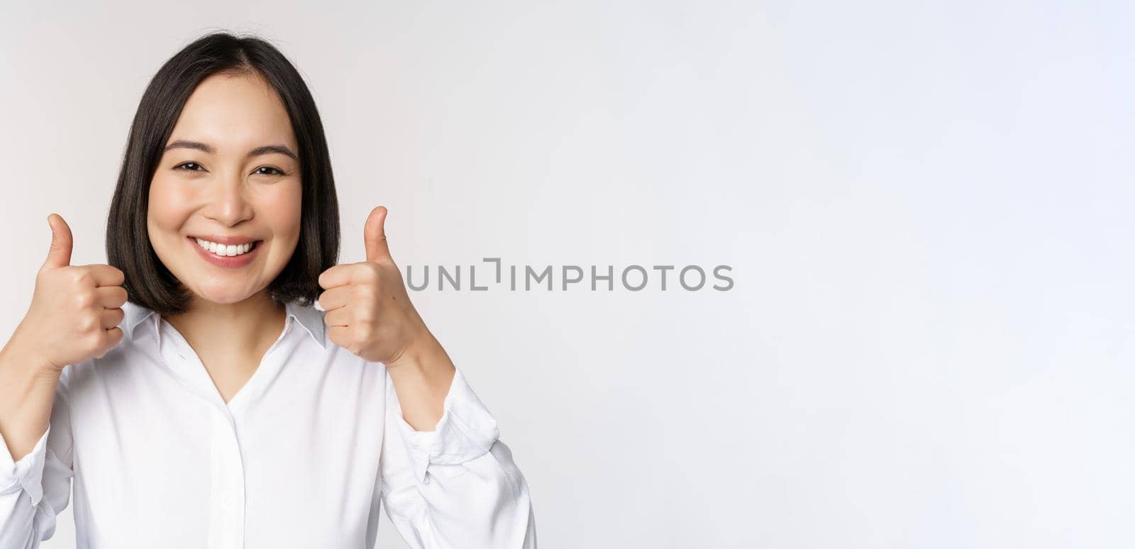 Cheerful asian woman face, showing thumbs up and smiling, pleased by something, recommending smth good, standing over white background.