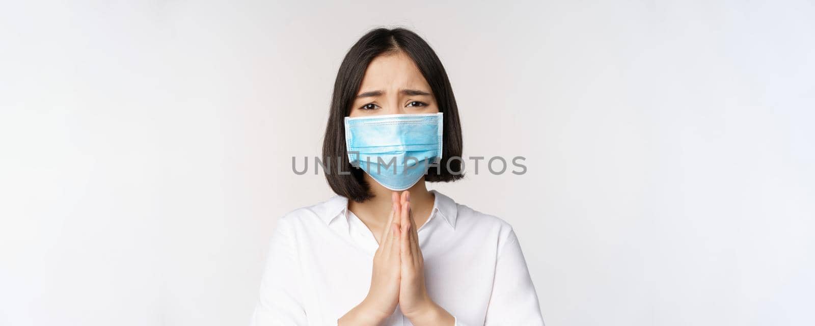 Portrait of asian woman in medical face mask from covid, begging, asking for help, say please, standing over white background by Benzoix