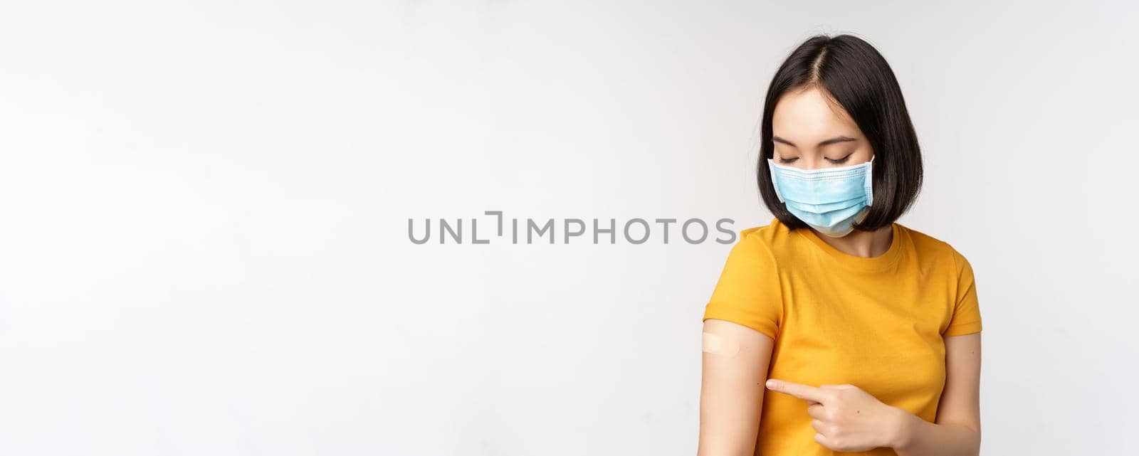 Covid-19, vaccination and healthcare concept. Cute asian girl in medical face mask, showing band aid after coronavirus vaccination, standing over white background.