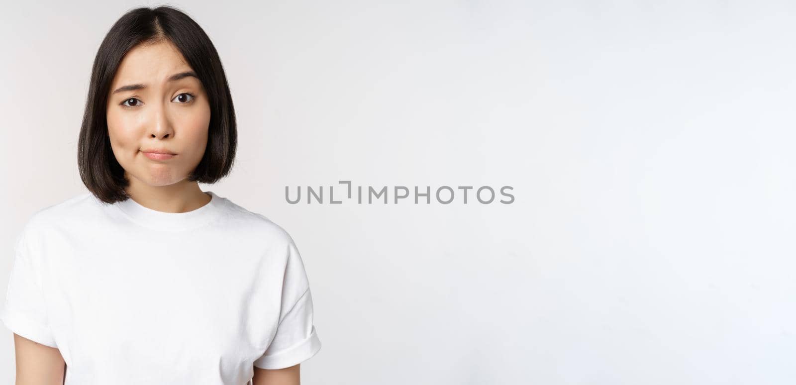 Image of young asian brunette woman in white t-shirt, smirk and look with doubt, hesitating, standing against white background by Benzoix