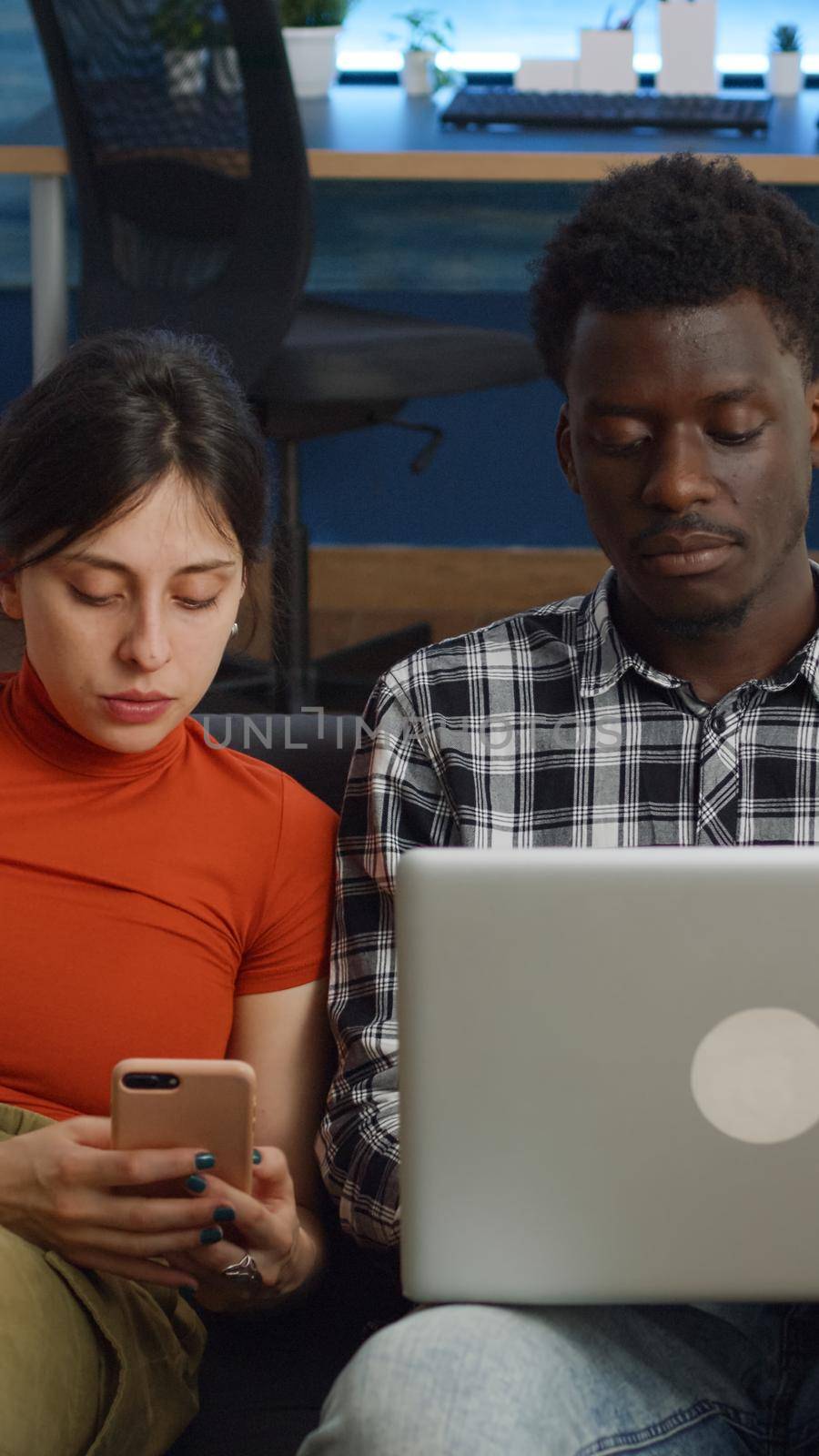Modern interracial couple with technology sitting on sofa together in living room. Mixed race married lovers using smartphone and laptop at home on couch. Multi ethnic family with gadgets