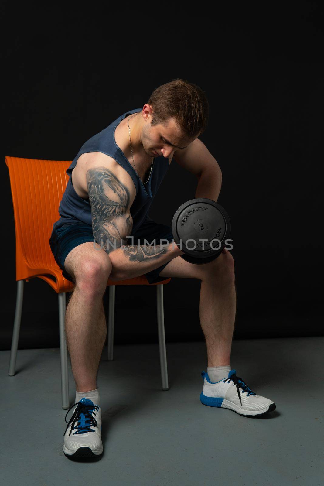 Man on black background sits on a chair rocking his biceps keeps dumbbells pumped up in fitness bodybuilding black, weight fitness athletic hand, lifestyle. Attractive sportive power, guy fit