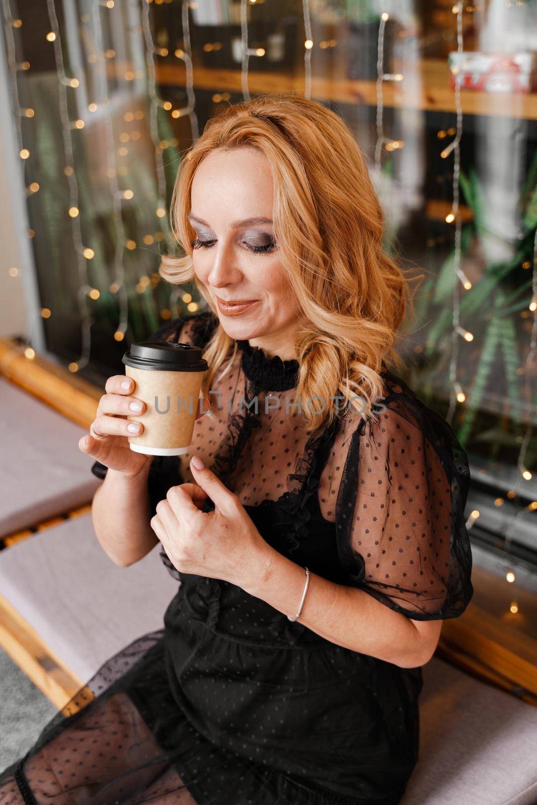 Young charming blonde with a cute smile and makeup while relaxing in a cafe. She is holding a cup of coffee in her hands. She is dressed in a black dress with transparent sleeves. by Matiunina