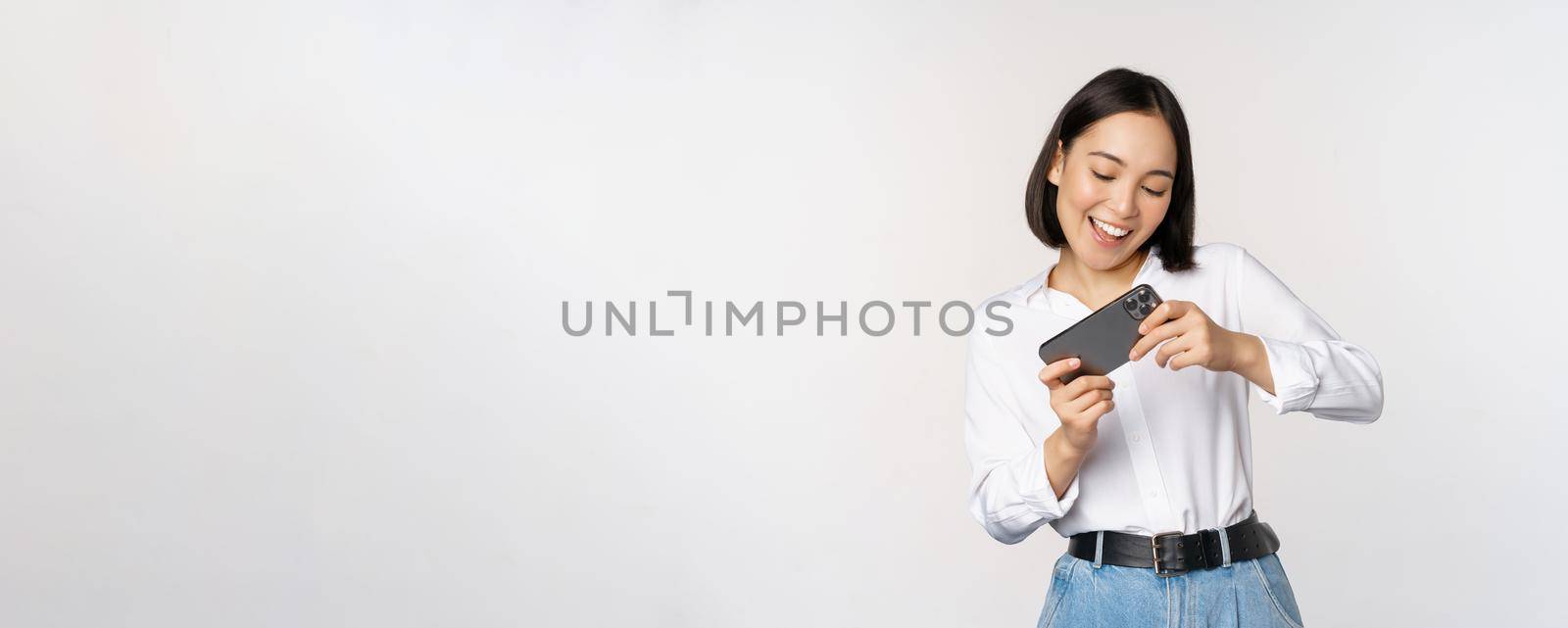 Young korean woman, asian girl playing mobile video game on smartphone, looking at horizontal phone screen, standing over white background.
