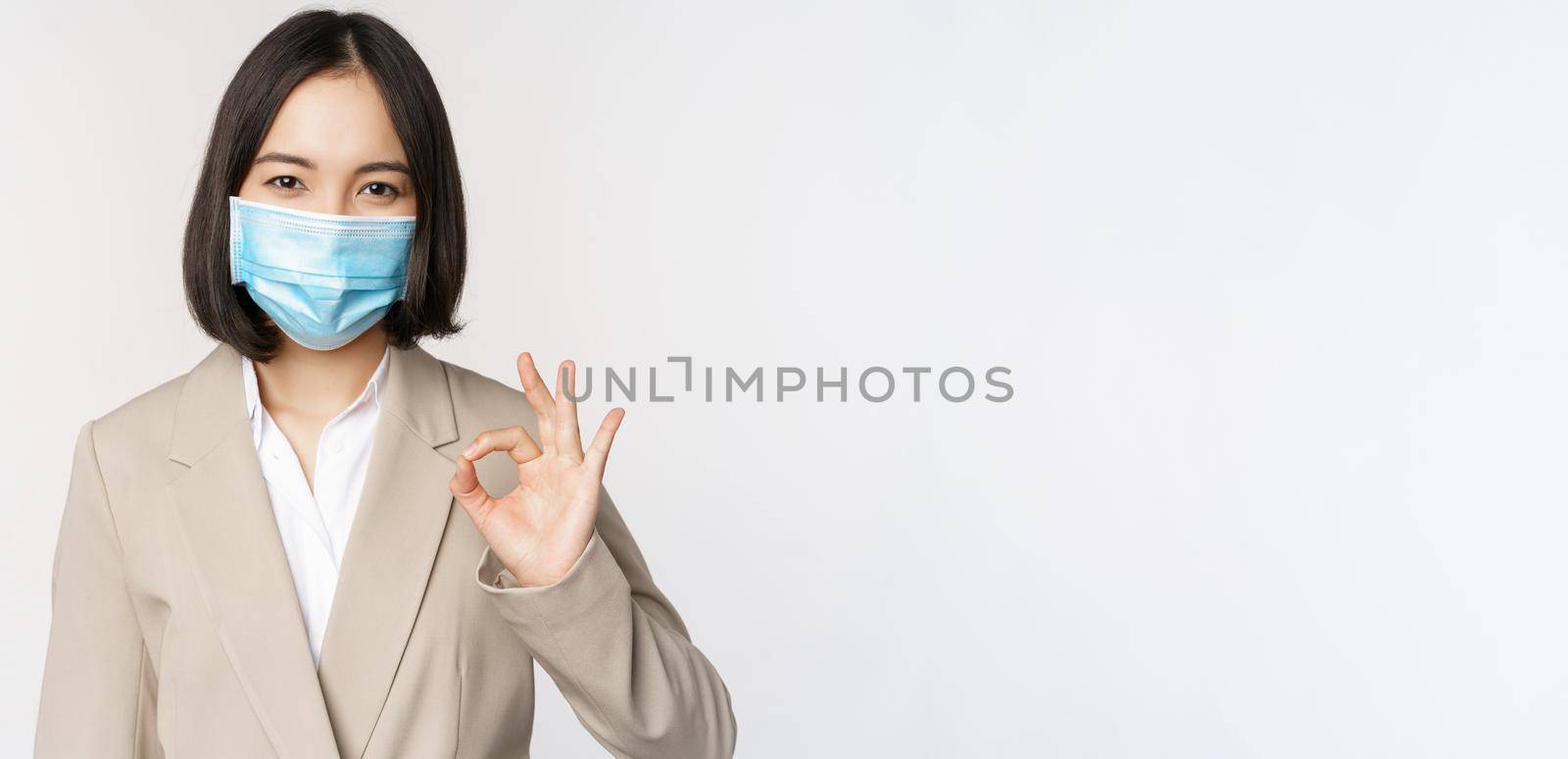 Coronavirus and workplace concept. Image of asian saleswoman, company worker in medical mask showing okay sign, smiling pleased, white background by Benzoix