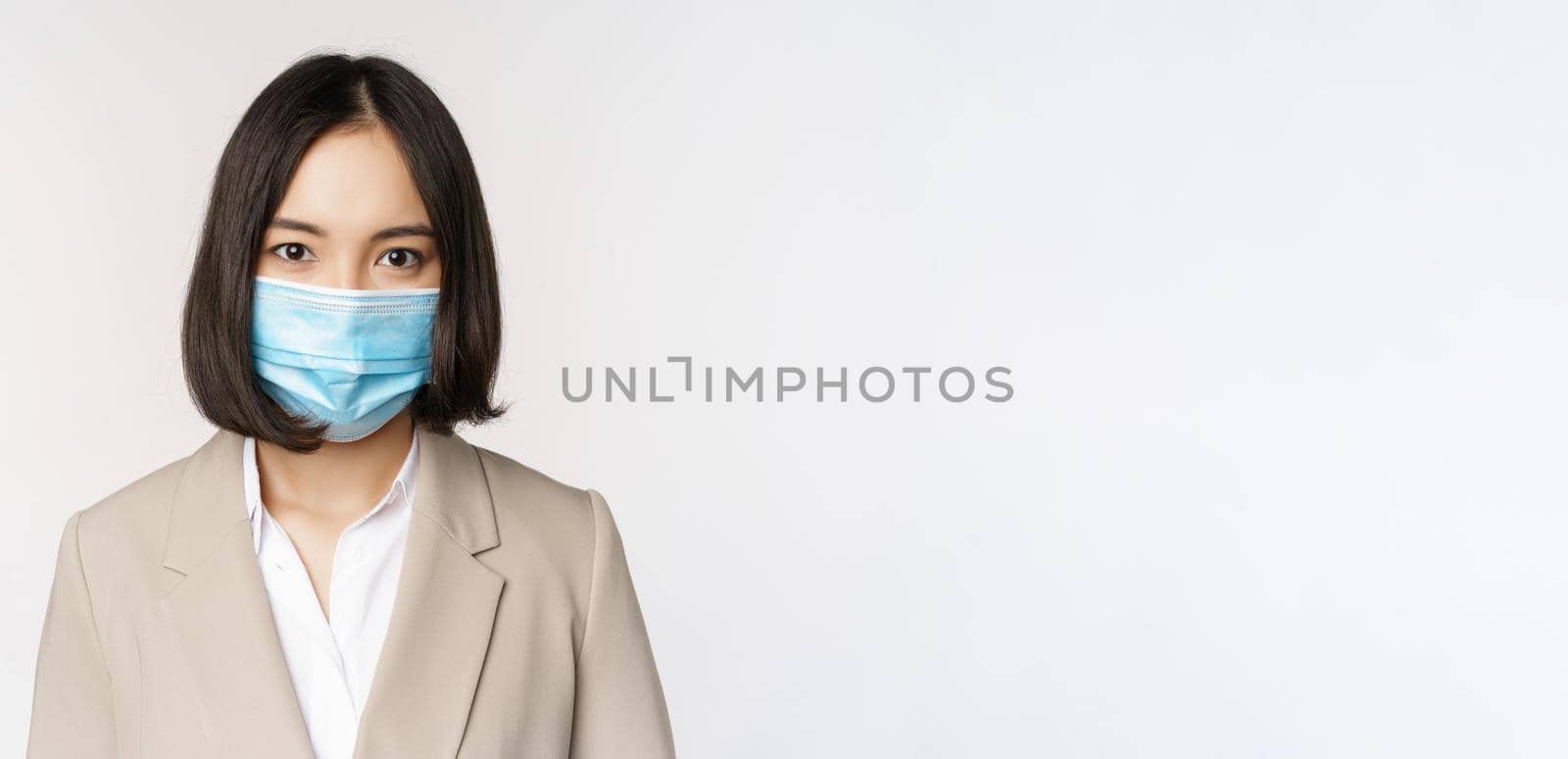 Coronavirus and workplace concept. Portrait of asian office worker, businesswoman in medical face mask looking at camera, white background by Benzoix