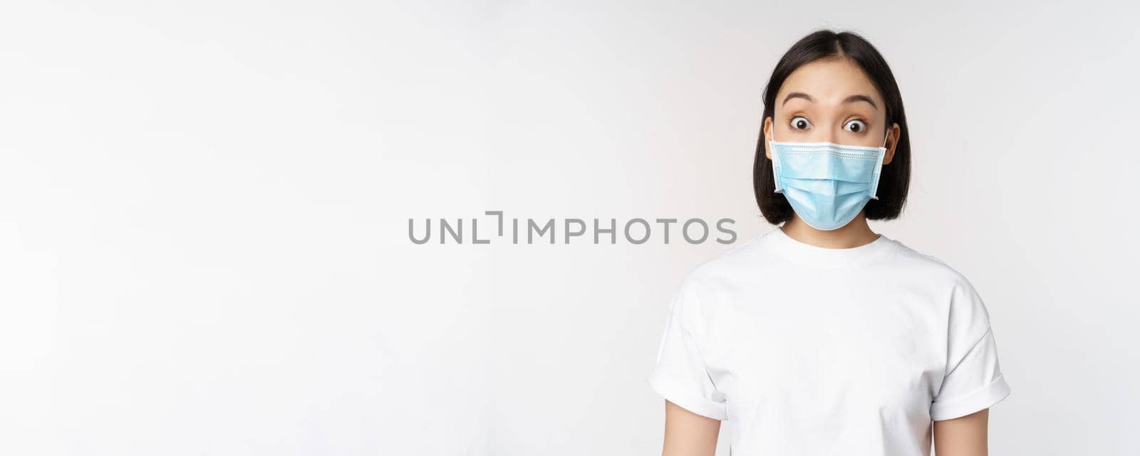 Health and covid pandemic concept. Image of korean woman in face medical mask looking surprised, reacting amazed, standing over white background.