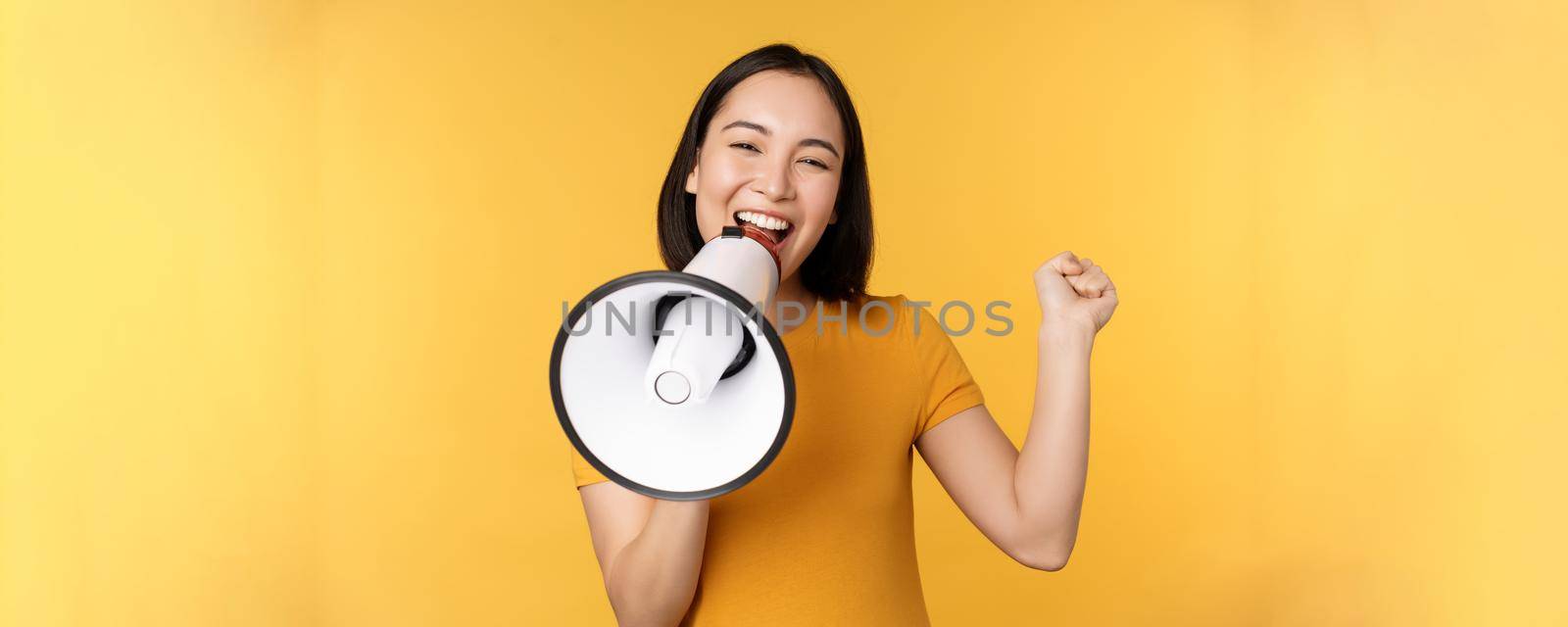 Smiling asian woman standing with megaphone, announcing smth, advertising product, standing over yellow background.