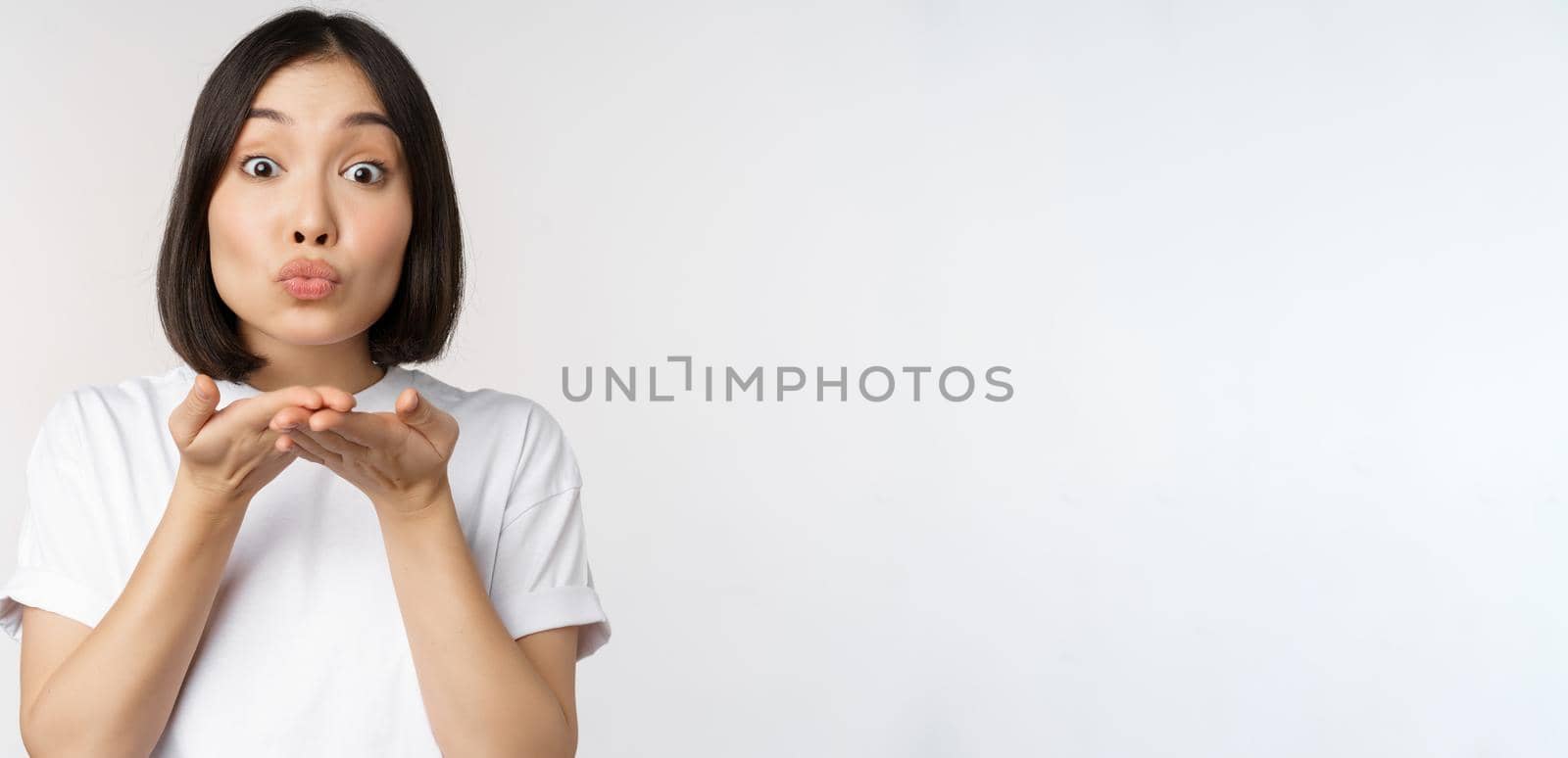 Lovely young asian woman, sending air kiss and looking coquettish at camera, standing in tshirt over white background.